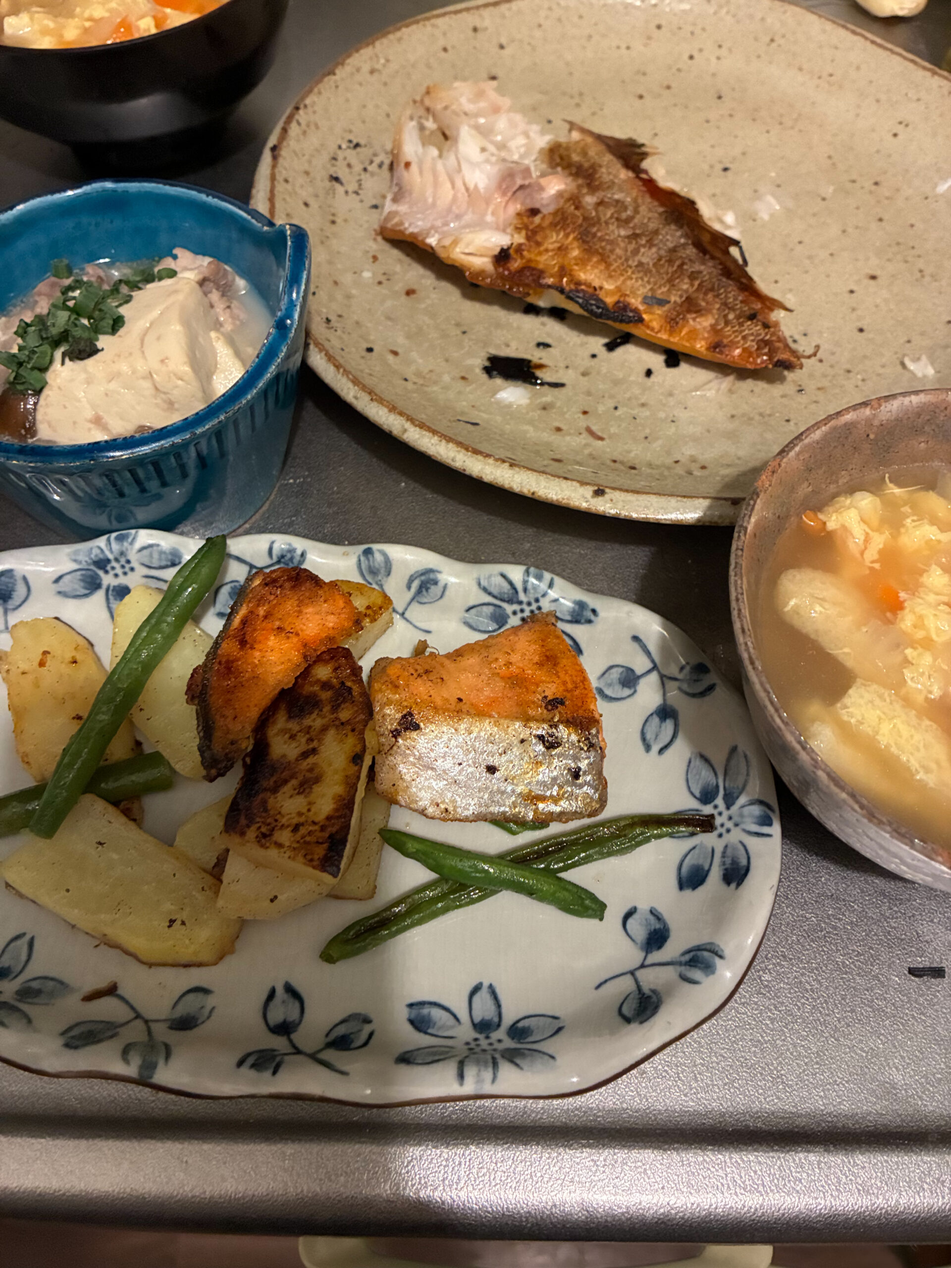 食卓に並んだ焼き魚の和定食。赤魚の干物、鮭とじゃがいもの焼き物、肉豆腐、かき玉汁などが並ぶ。