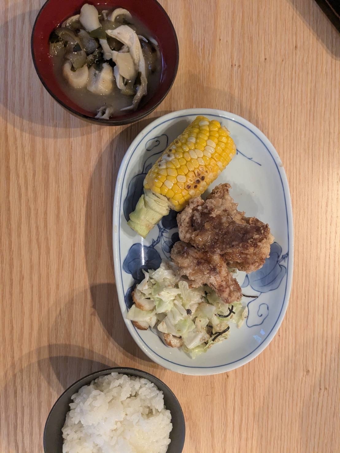 木目のテーブルに置かれた、とうもろこしと唐揚げの和定食。白い楕円形の皿に焼きとうもろこし、唐揚げ、キャベツの和え物が盛り付けられ、奥には味噌汁、手前にはご飯が添えられている。