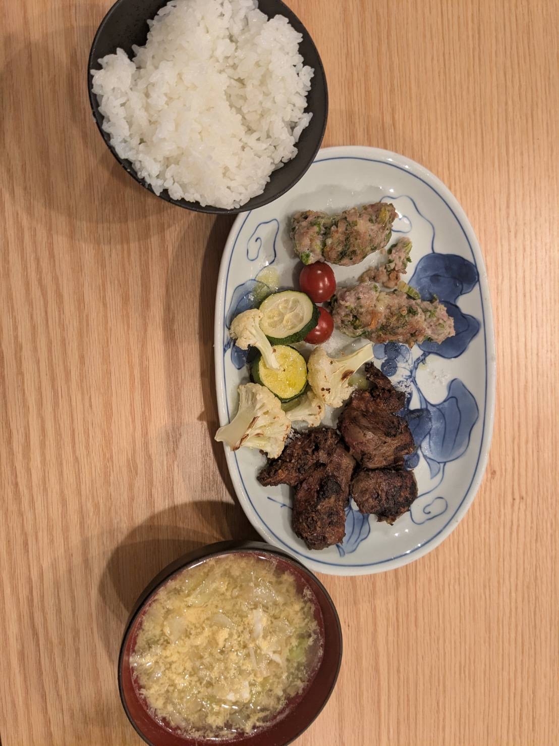 肉と野菜のおかずが乗ったお皿と、白米、卵とじの味噌汁が並んだ食卓の写真