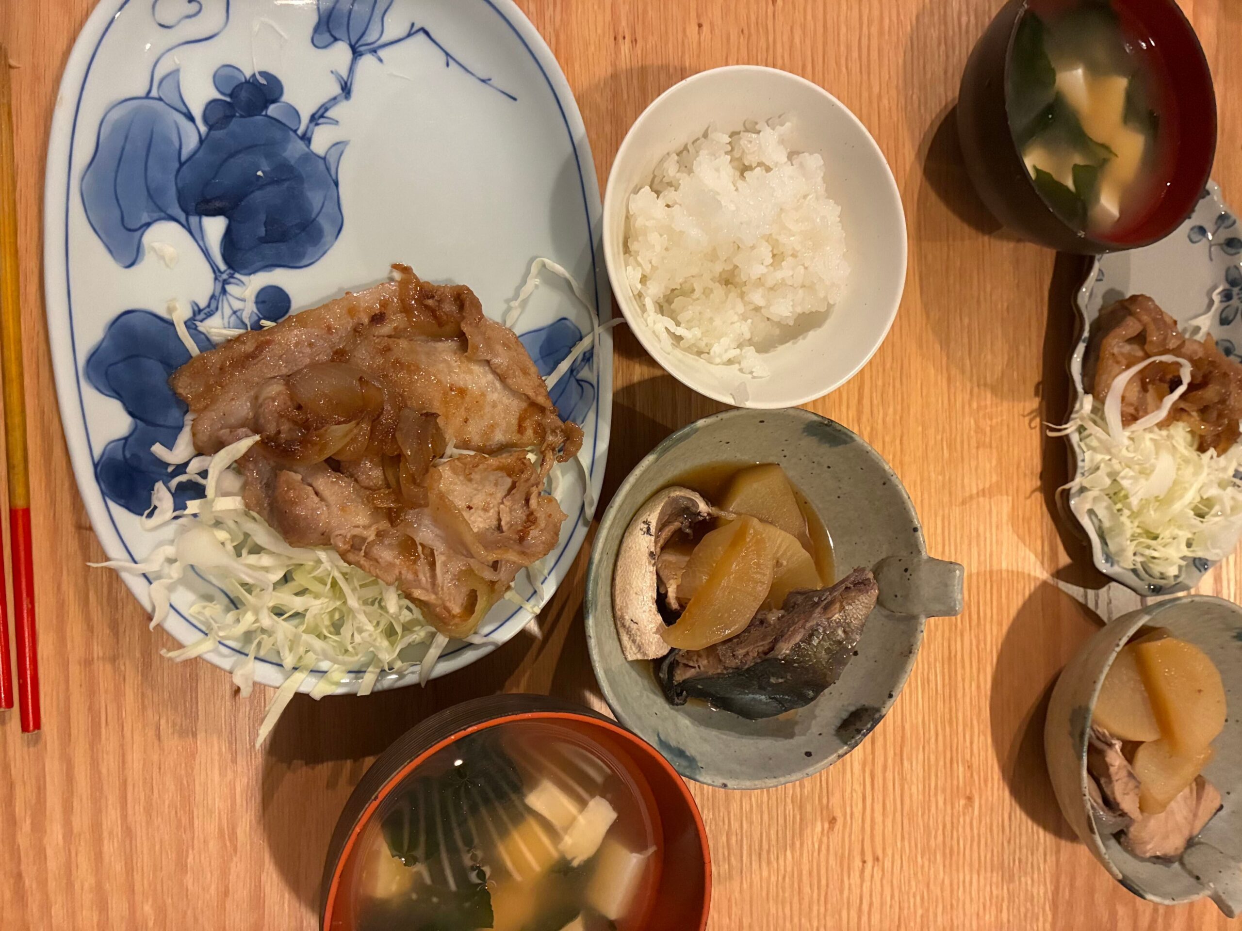 豚の生姜焼き定食が並んだ食卓。大皿の生姜焼き、ごはん、味噌汁、ぶりの煮付けが木目のテーブルに置かれている。